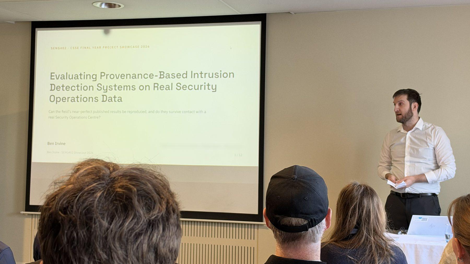 Ben Irvine speaks to a seated audience beside a projected title slide that reads “Evaluating Provenance-Based Intrusion Detection Systems on Real Security Operations Data”.